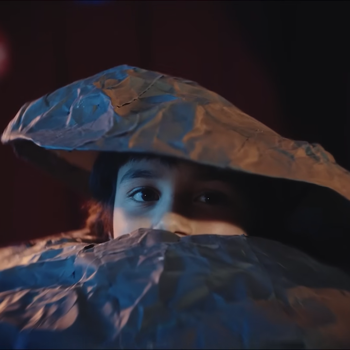 Child peeking over crumpled blue paper hats, eyes wide in curiosity under a makeshift cover.